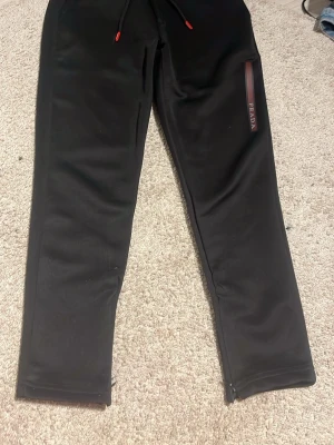 Svarta mjukisbyxor från Prada - Snygga svarta mjukisbyxor från Prada med dragsko i midjan och röda detaljer på snöret. Byxorna har en smal passform och dragkedjor vid bensluten. Materialet ser mjukt och bekvämt ut, perfekt för en chill look.