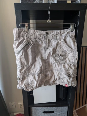 Beige kortkjol med fickor och dragkedja - Snygg beige kortkjol med flera fickor, dragkedjedetaljer och knappstängning. Kjolen har en avslappnad look med synliga sömmar och passar perfekt till en casual stil. Praktiska fickor både fram och bak ger en cool utility-vibe.