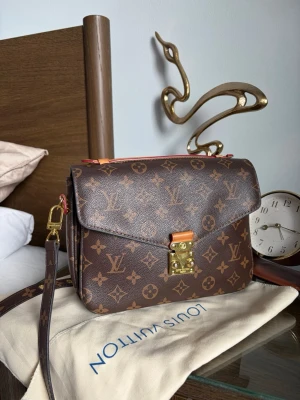 Louis Vuitton Väska - Säljer en brun Louis Vuitton Pochette Métis handväska i klassiskt monogrammönster. Väskan har guldiga metalldetaljer, justerbar axelrem och flera fack inuti. 