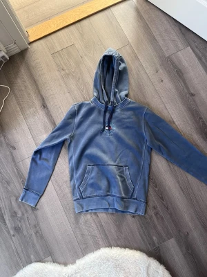 Blå hoodie från Tommy Jeans - Säljer en blå hoodie från Tommy Jeans med broderad logga och flagga på bröstet. Hoodien har känguruficka, snörning i huvan och långa ärmar. Perfekt för en chill och avslappnad stil.
