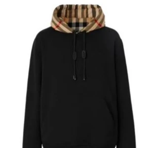 Svart hoodie med Burberry-rutig huva - Snygg svart hoodie från Burberry med klassisk beige, röd och svart rutig huva. Hoodien har dragsko vid huvan och en avslappnad passform. Perfekt för dig som vill ha en stilren look med en touch av lyx.