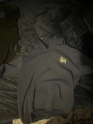 Svart Stüssy hoodie med logga - Säljer en svart hoodie från Stüssy med deras ikoniska logga i gult tryck på bröstet. Hoodien har en klassisk känguruficka framtill och huva med dragsko. Perfekt för dig som gillar streetwear och vill ha en stilren men ändå statement tröja.