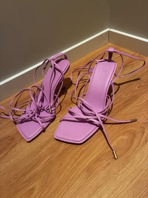 Rosa strappy sandaler  - Säljer ett par trendiga rosa sandaler med fyrkantig tå och tunna remmar som snöras runt benet. Sandalerna har en stilren design med guldfärgade detaljer på snörändarna. Perfekta för att lyfta din sommarlook.