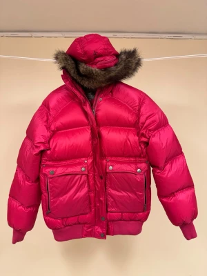 Rosa/röd dunjacka från Cross Sweden - Snygg röd dunjacka med stor pälskantad huva och två stora framfickor med knappar. Jackan har ribbade muddar vid ärmslut och nederkant samt en glansig finish. Avtagbar både päls och hela huvan. Perfekt för kalla vinterdagar och riktigt varm.