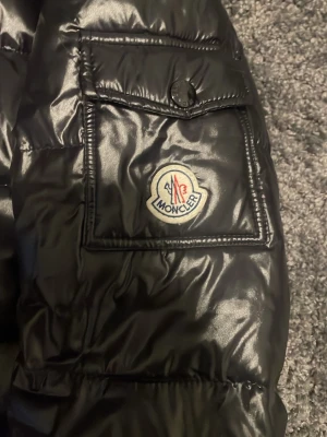 Svart dunjacka från Moncler - Snygg svart dunjacka från Moncler med glansig finish, huva och dragkedja framtill. Jackan har fickor med dragkedja och en extra ficka med knapp på ärmen där Moncler-loggan sitter. Perfekt för kalla vinterdagar.