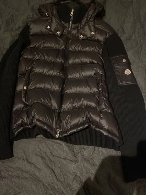 Svart dunväst från Moncler - Säljer en svart dunväst från Moncler med stickade ärmar och huva. Västen har quiltad front i blank polyester, dragkedja och fickor med tryckknappar. Klassisk Moncler-logga på ärmen och ribbade detaljer för extra stilpoäng. Storlek XXL, men de som vet om Moncler storlekarna vet att dessa modeller sitter mycket mindre, skulle säga att den passar folk med storlekarna Large Medium, Large, och XL!