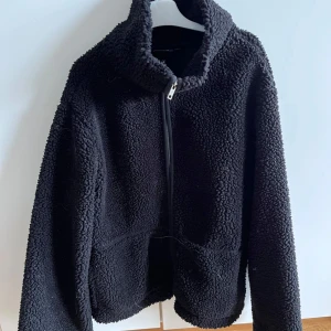 Svart teddyjacka från lager 157, stl S - Mysig svart teddyjacka från lager 157 i storlek S. Jackan har hög krage, dragkedja framtill och är tillverkad i fluffigt teddy-material som håller dig varm. Perfekt för kyliga dagar och enkel att matcha med olika outfits.
