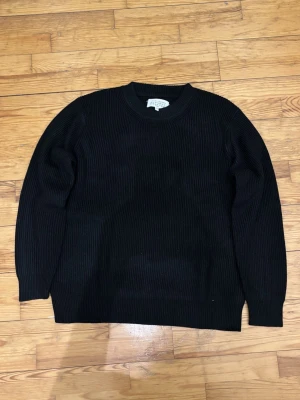 Svart stickad tröja Maison Margiela - Säljer en svart, ribbstickad tröja från Maison Margiela. Klassisk rund halsringning och långa ärmar. Tröjan har Margielas ikoniska vita stygn i nacken och är gjord i ett tjockare stickat material som passar perfekt till kyligare dagar.