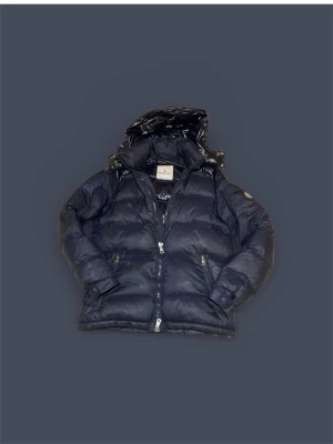 Mörkblå Moncler pufferjacka med huva - Säljer en mörkblå pufferjacka från Moncler med avtagbar huva och två fickor med dragkedja. Jackan har klassisk Moncler-logga på ärmen och är tillverkad i glansig polyester med dunfyllning. Perfekt för kalla dagar och riktigt snygg till streetwear.