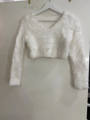 Vit fluffig croppad tröja - Säljer en vit fluffig croppad tröja med lång ärm och v-ringad hals. Tröjan har en mysig pälsliknande yta som ger en cool och trendig look. Perfekt för dig som gillar att sticka ut och vill ha något extra mjukt och skönt.