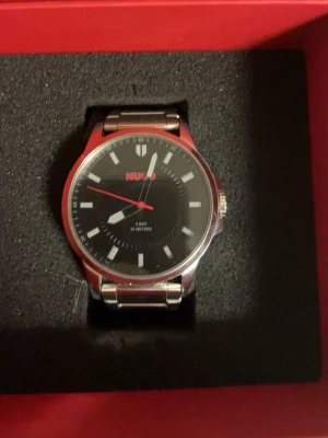 HUGO  svart klocka med metallarmband - Snygg klocka från HUGO  med svart urtavla och röda detaljer. Klockan har ett stilrent metallarmband i silverfärg och minimalistiska markeringar. Urtavlan är rund och har en modern look med HUGO-loggan i rött. Perfekt accessoar för dig som gillar stilrena detaljer. Du får med extra metallarmband. Lite repor på armbandet. Knappt använd. Ord pris 1800