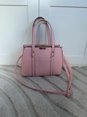 Ljusrosa axelväska med gulddetaljer - Säljer en ljusrosa axelväska i syntetmaterial med både handtag och justerbar axelrem. Väskan har flera fack, dragkedja upptill och guldfärgade metalldetaljer. Insidan är beige och har smarta innerfickor för organisering. Perfekt för dig som vill ha en stilren och söt väska.