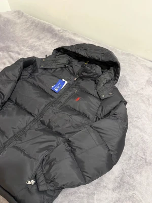 Ralph Lauren jacka  - Säljer en svart pufferjacka från Polo Ralph Lauren med huva och dragkedja framtill. Jackan har en röd broderad logga på bröstet, flera fickor med dragkedja och en klassisk pufferlook. Perfekt för kalla dagar och riktigt snygg till streetwear.