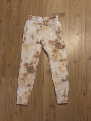 Bruna tie-dye mjukisbyxor från Lindex - Säljer ett par mjukisbyxor från Lindex i storlek 146/152 med brun och beige tie-dye effekt. Byxorna har resår i midjan och vid bensluten samt snörning i midjan. Insidan är mjuk och fleece-liknande för extra komfort.