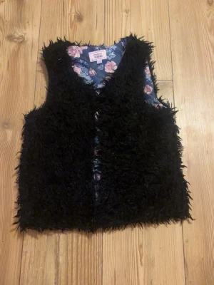 Svart pälsväst från Mywear Young - Säljer en fluffig svart pälsväst från Mywear Young. Västen har ett mjukt, lurvigt yttre och ett blommigt foder i blått och rosa toner. Perfekt för dig som vill addera lite extra stil till din outfit.