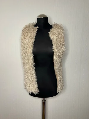 Beige fluffig pälsväst Appletree Boutique - Säljer en supermjuk och fluffig beige pälsväst från Appletree Boutique. Västen har en öppen design utan knäppning och är fodrad med glansigt tyg på insidan. Perfekt för att ge din outfit en extra touch av stil.