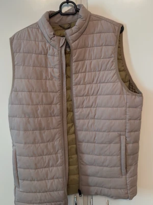 Beige quiltad dunväst Jack & Jones - Säljer en beige quiltad dunväst från Jack & Jones i storlek S. Västen har ståkrage, dragkedja framtill och två sidofickor. Perfekt att slänga över en hoodie för extra värme. Enkel och clean design som funkar till det mesta.