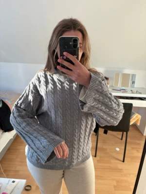 Grå kabelstickad tröja med vida ärmar - Mysig grå kabelstickad tröja med breda, långa ärmar och rund hals. Tröjan har ett klassiskt flätmönster och är perfekt för kyligare dagar. Passformen är loose och materialet känns mjukt och värmande. Den passar även mamma som är en L så den är rätt pösig. Knappt använd! 
