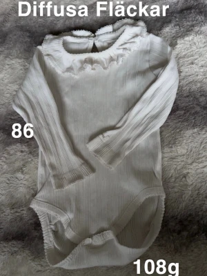 Vit ribbad body med volangkrage - Söt vit body med långa ärmar och volangkrage. Ribbad struktur och knäppning i grenen för enkel påklädning. Perfekt för lager-på-lager och passar till många olika outfits.🌸 Diffusa fläckar. Storlek 86