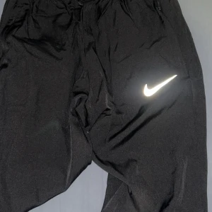 Svarta mjukisbyxor från Nike - Svarta mjukisbyxor från Nike med klassisk vit logga på benet. Byxorna har resår i midjan och är tillverkade i mjukt material som passar perfekt för chill eller träning. Snygg och enkel design med avslappnad passform.