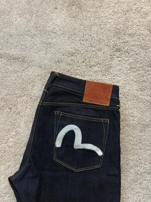 Evisu jeans - Klassiska Evisu-jeans i mörk, rå denim och en femficksdesign med detaljer i kopparfärgade nitar. Tveka inte att höra av er vid frågor!