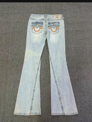 True Religion Joey Low Rise Flare Jeans - Säljer ett par ljusblå True Religion Joey Low Rise Flare jeans med ikoniska broderade fickor bak och orange detaljer. Byxorna har låg midja, utsvängda ben och klassisk knapp med logga. Perfekta för dig som gillar retrovibbar och statement-jeans.
