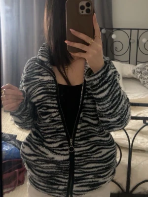 Zebramönstrad fleecekofta med dragkedja - Säljer en fluffig och mysig kofta i zebramönster med svart och vit färg. Koftan har en hög krage och hel dragkedja framtill med rund metallring. Materialet är mjuk fleece och passformen är loose, perfekt för kyliga dagar.