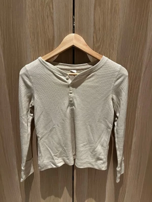 Beige ribbad långärmad topp - Säljer en beige långärmad topp i ribbad bomull med fyra knappar framtill. Toppen har en enkel och clean look med rundad halsringning och smal passform. Perfekt att styla med jeans eller kjol för en chill vibe.