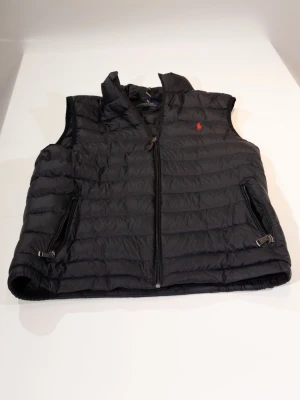 Svart dunväst från Polo Ralph  Lauren - Säljer en svart dunväst från Polo Ralph Lauren med klassisk röd logga på bröstet. Västen har quiltad design, hög krage och två fickor med dragkedja. Perfekt att slänga över en hoodie eller tröja för extra värme. Stängs med dragkedja framtill.