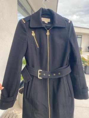 Svart ullkappa från Michael Kors - Snygg svart ullkappa från Michael Kors med guldfärgade dragkedjor och detaljer. Kappan har ett brett bälte i midjan, långa ärmar och klassisk krage. Perfekt för dig som vill ha en stilren och lyxig look under höst och vinter.