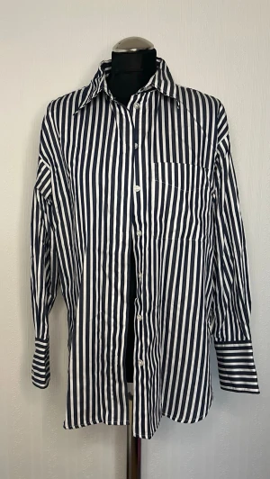Randig skjorta från H&M i bomull - Snygg skjorta från H&M med marinblå och vita vertikala ränder. Klassisk krage, bröstficka och knäppning framtill. Tillverkad i mjuk bomull och har långa ärmar. Perfekt för en clean och stilren look.