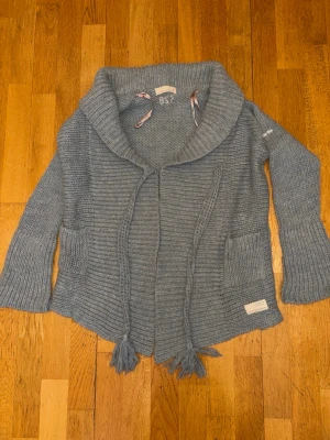 Grå stickad kofta med snörning - Mysig grå stickad kofta från Odd Molly med bred krage, två stora fickor framtill och dekorativa snören med tofsar. Koftan har långärmad design och ribbade detaljer som ger en avslappnad och cool look.