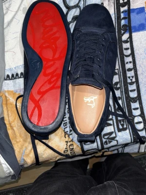 Mörkblå mocka sneakers från Louboutin - Säljer ett par mörkblå sneakers från Christian Louboutin i mjuk mocka med klassisk röd sula och signaturbrodyr på hälen. Skorna har rund tå, snörning och en stilren design som sticker ut med den ikoniska röda undersidan.