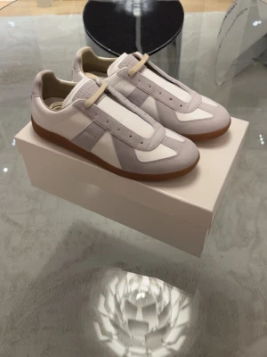 Maison Margiela sneakers vit/grå - Säljer ett par stilrena Maison Margiela sneakers i vitt och ljusgrått skinn och mocka. Klassisk låg modell med gummisula och rund tå. Snygga detaljer i mocka och diskret logga på plösen. Perfekta till jeans eller dressade byxor.