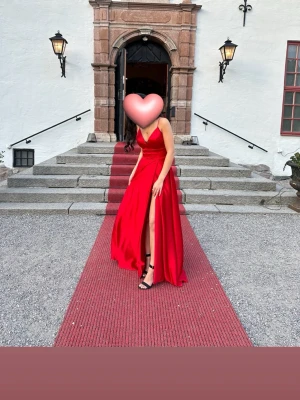 Röd långklänning med slits och snörning - Säljer en röd långklänning i glansigt material med smala axelband, djup v-ringning och hög slits framtill. Klänningen har snygg snörning i ryggen som ger en extra detalj. Perfekt för bal eller student!