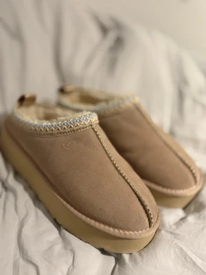Beige UGG mockatofflor med fårfoder - Säljer ett par beige UGG tofflor i mocka med fluffigt fårfoder och grov sula. Dekorativt mönster i blått och beige runt öppningen. Perfekta för kalla dagar och riktigt sköna att ha på sig hemma.
