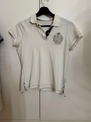 Lauren - Polo Jeans Co. - Säljer en fantastisk vit pikétröja från Ralph Lauren - Polo Jeans Co. linjen. Detta ar inte din vanliga piké; den har ett stort, detaljerat emblem på bröstet med broderier och små pärlor som ger en lyxig vintagekänsla. Modellen är figurnära med snygga kontrastdetaljer i mörkblått vid knappslået och dekorativa sömmar. Perfekt för en Y2K-inspirerad look! • Skick: Mycket gott vintageskick. • Detaljer: Tryckknappar, broderat emblem, unika sömmar. • Stil: Y2K, Vintage, Streetwear.