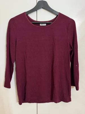 Vinröd topp med trekvartsärm från Dea - Klassisk och mjuk topp i en djup vinröd färg från märket Dea. Denna modell har en smickrande båthals med en elegant satinkant längs ringningen, vilket ger den där lilla extra touchen. Toppen har trekvartsärm, vilket gör den perfekt för både vardag och lager-på-lager under en kavaj. Ett mångsidigt basplagg som aldrig går ur tiden! • Märke: Dea • Storlek: M • Färg: Vinröd / Burgundy • Skick: Fint skick