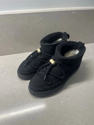 Svarta Inuikii boots  - Säljer ett par svarta Inuikii boots med mjuk pälsfodring och grova snören med gulddetaljer. Skorna är i mocka och har en platt sula samt rund tå. Perfekta för kalla dagar och ger en riktigt mysig vinterlook.