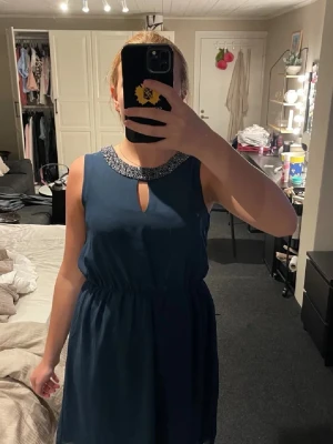 Mörkblå ärmlös klänning med pärldetaljer - Supersnygg mörkblå klänning med pärlbroderad krage och liten öppning vid halsen. Klänningen är ärmlös, har resår i midjan och faller i en mjuk, luftig form. Perfekt för dig som vill ha något elegant och bekvämt.