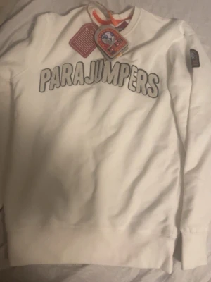 Vit sweatshirt från Parajumpers - Säljer en clean vit sweatshirt från Parajumpers med stor logga broderad över bröstet och klassisk patch på ärmen. Tröjan har rund halsringning och långa ärmar. Perfekt för dig som gillar stilrena och sportiga plagg. Öppen för byte 