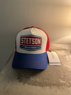 Stetson röd, vit och blå truckerkeps - Säljer en riktigt snygg truckerkeps från Stetson med vit front, röd mesh och blå skärm. Kepsen har broderad Stetson-logga framtill och justerbar snapback. Perfekt för dig som vill sticka ut med klassisk amerikansk stil.