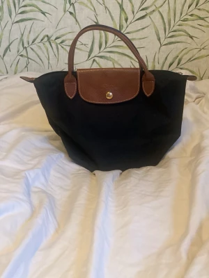 Svart Longchamp Le Pliage - Klassisk svart handväska från Longchamp Le Pliage ”Deposé” med bruna skinnhandtag och lock. Den är i perfekt skick och jag fick den som julklapp! Väskan har guldfärgade detaljer och dragkedja upptill. Rymlig insida med en liten innerficka. Perfekt storlek för vardagsgrejer och enkel att bära med sig. Skriv för mer bilder eller info!💞💞