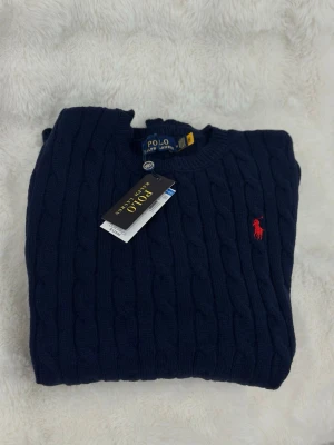 Marinblå kabelstickad tröja Polo Ralph Lauren - Kabelstickad marinblå tröja från Polo Ralph Lauren med klassisk röd logga broderad på bröstet. Rund halsringning och långa ärmar. Tröjan är gjord i mjuk bomull och har ribbade muddar vid ärmslut och nederkant. Perfekt för en clean och stilren look.