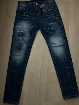 Dsquared2 blå jeans med slitningar - Säljer ett par mörkblå jeans från Dsquared2 med coola slitningar och vita färgstänk. Jeansen har klassisk femficksdesign, rak passform och snyggt tvättad look. Materialet är robust denim och de har en röd Dsquared2-logga vid gylfen.