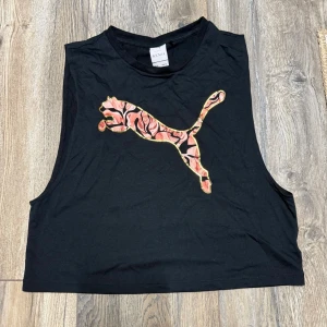 Svart croppad linnetopp Puma Kenza - Säljer en svart croppad linnetopp från Puma x Kenza med ett stort färgglatt Puma-tryck framtill. Toppen är ärmlös med breda axelband och har en avslappnad passform. Perfekt till jeans eller shorts för en sportig vibe.