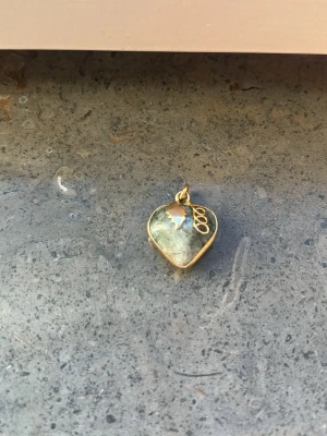 Hjärthänge i grön sten och guld - Unikt hjärthänge med grönskimrande sten infattad i guldfärgad metall. Hänget har ett dekorativt blad och spiral i metall på framsidan. Perfekt som personlig detalj till halsband eller armband.