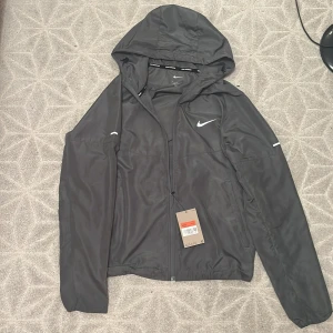 Nike miller  - Grå vindjacka från Nike med huva och dragkedja framtill. Jackan har diskret Nike-logga på bröstet, elastiska muddar och två sidofickor. Pris kan diskuteras.