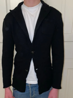 John Henric Sticksd Merino Kavaj - Svart stickad kavaj med klassisk krage och två knappar framtill. Jackan har tre fickor, varav en bröstficka, och är långärmad med en normal passform. Perfekt för dig som gillar en chill och stilren look. Modellen är cirka 183 och väger 70kg. Kavajen är M men skulle snarare säga att den sitter som S.