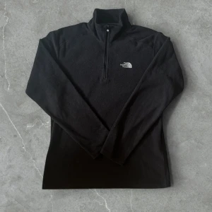 Svart fleece-tröja från The North Face - Svart fleece-tröja från The North Face med halv dragkedja och hög krage. Perfekt för kyliga dagar, med broderad logga på bröstet. Långärmad och mjuk. Storlek S men passar M
